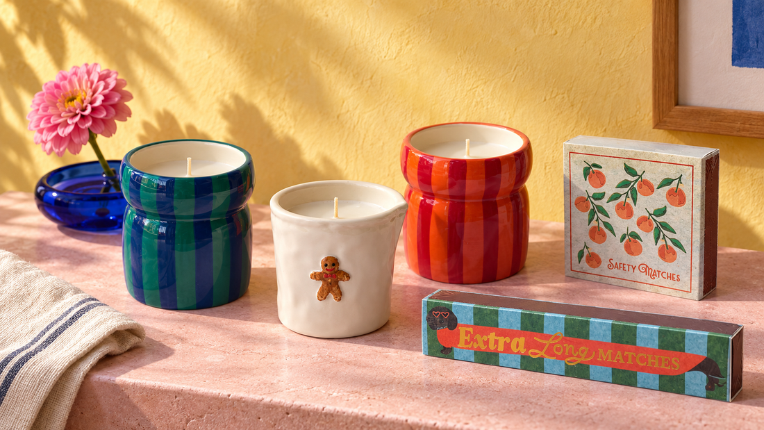 Três velas coloridas e duas caixas de fósforos sobre uma mesa, junto a uma jarra azul com uma flor cor-de-rosa.